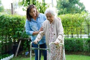 Caregiver help Asian elderly woman disability patient walk with walker in park, medical concept. photo