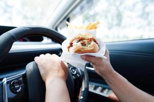 Asian woman driver hold and eat hamburger and french fries in car, dangerous and risk an accident. photo