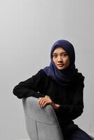 Serious Asian hijab woman in casual shirt sitting on chair, posing with various hand gesture. Isolated studio portrait on gray background. photo