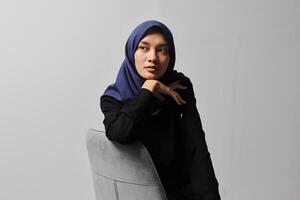 Serious Asian hijab woman in casual shirt sitting on chair, posing with various hand gesture. Isolated studio portrait on gray background. photo
