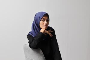 Serious Asian hijab woman in casual shirt sitting on chair, posing with various hand gesture. Isolated studio portrait on gray background. photo