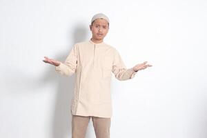 Portrait of confused Asian muslim man in koko shirt with skullcap spreading his hands sideways and holding two things, demonstrate products. Isolated image on white background photo