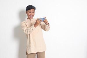 Portrait of young excited Asian muslim man in koko shirt holding mobile phone and playing games on his smartphone. Isolated image on white background photo