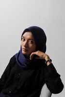 Serious Asian hijab woman in casual shirt sitting on chair, posing with various hand gesture. Isolated studio portrait on gray background. photo