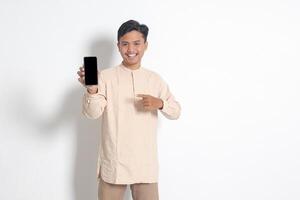 Portrait of young excited Asian muslim man in koko shirt showing blank screen mobile phone mockup while pointing and presenting product. Social media concept. Isolated image on white background photo