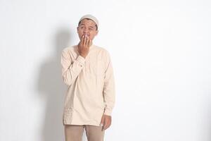 Portrait of overworked Asian muslim man in koko shirt with skullcap yawning with hand covering his mouth. Sleep deprivation concept. Isolated image on white background photo