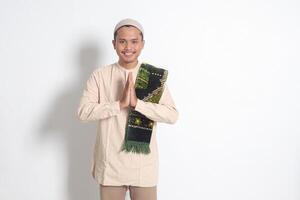 Portrait of attractive Asian muslim man in koko shirt with prayer mat showing apologize and welcome hand gesture. Apology during eid mubarak. Isolated image on white background photo