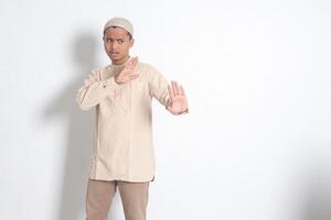 Portrait of unpleasant Asian muslim man in koko shirt with skullcap forming a hand gesture to avoid something or bad things. Isolated image on white background photo