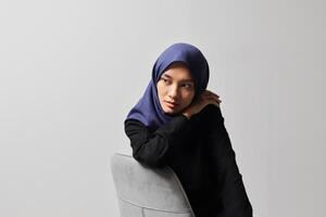 Serious Asian hijab woman in casual shirt sitting on chair, posing with various hand gesture. Isolated studio portrait on gray background. photo