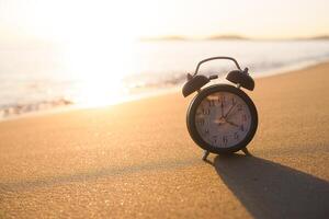 negro alarma reloj en el playa en el puesta de sol tiempo. el concepto acerca de hora a verano, viajar, vacaciones y relajación foto