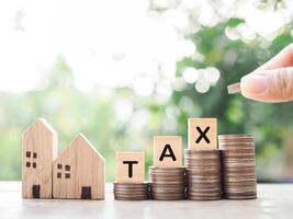 Close up of hand putting coins into stack of coins, Miniature house and wooden blocks with the word TAX. The concept of payment tax for house, Property investment, House mortgage, Real estate. photo