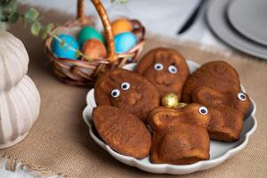 Close-up of funny muffins shaped as bunnies and eggs with eyes, multi colored Easter eggs in wicker basket on served table photo