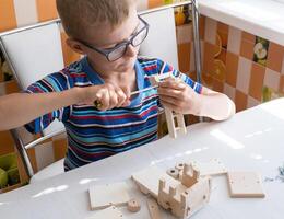 Portrait of a 7-8 year old boy with a screwdriver, carefully assembling a wooden car, sitting at the kitchen table. hands close up photo