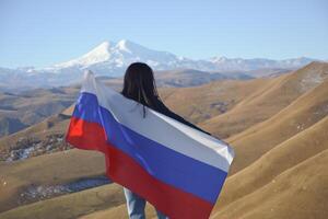 A young brunette woman stands against the backdrop of the snow-capped Mount Elbrus, looking at the beauty of the mountains, holding a Russian flag in her hands. Tricolor against the backdrop of snow photo
