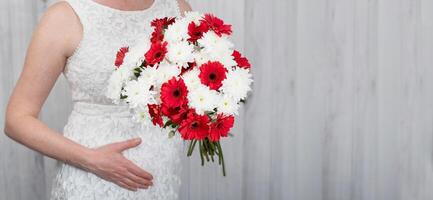A pregnant woman over 40 in a white dress holds her belly with her hand, a bouquet of white and red flowers in the other hand, copy space photo