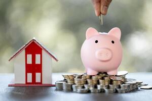 Model of house with female hand putting coin in piggy bank on blurred background. Money saving or investing for home loan or real estate. Investment property, Saving money for retirement concept. photo