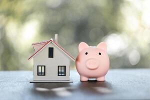 Model of house with piggy bank on table on blurred background. Money saving or investing for home loan or real estate. Investment property, Saving money for retirement concept. photo