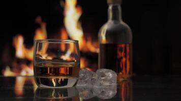 Glass and bottle of whiskey, scotch or bourbon stands on a table against background of a fireplace with a flame. Alcoholic drink and home comfort concept. video