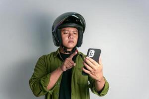 adulto asiático hombre confuso cuando mirando a su teléfono de mano mientras vistiendo motocicleta casco foto