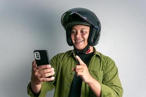 adulto asiático hombre sonriente cuando mirando a su teléfono de mano mientras vistiendo motocicleta casco foto