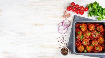Homemade meatballs with tomato sauce and spices served in black pan on white wooden background photo