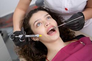 retrato de joven sonriente mujer sentado en estomatología clínica silla y del doctor manos con dental boca espejo y dental explorador herramientas preparando a examinar dientes condición. foto