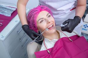 retrato de joven sonriente mujer sentado en estomatología clínica silla y del doctor manos con dental boca espejo y dental explorador herramientas preparando a examinar dientes condición. foto