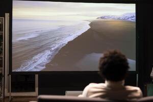 hombre relajante en sofá acecho clima cambio documental con hermosa cinematografía en moderno vídeo proyector. persona disfrutando paisaje disparos de mar playa durante amanecer en vivo habitación foto