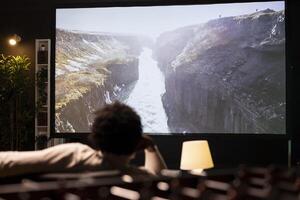 Homeowner chilling on couch in apartment watching nature documentary with breathtaking cinematography on huge TV screen. Man enjoying scenery shots on video on demand television channel photo