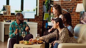 Multiracial group of people throwing birthday party for their friend, drinking alcohol and eating from charcuterie board. Merry guests laughing in modern living room after gifting host presents photo