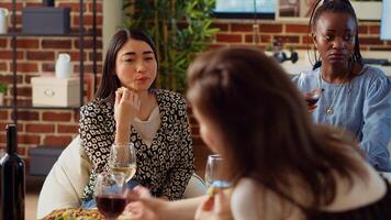 Asian woman at apartment party paying attention to interesting story shared by friend while eating gourmet cheese from charcuterie platter. BIPOC guest listening to group of people chatting photo