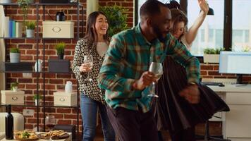 Multiracial group of friends dancing at apartment party, celebrating their friendship during annual reunion. Multiethnic colleagues doing energetic dance moves and laughing in cozy living room photo