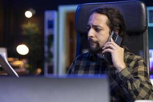 Serious man looking at documents while talking on cell phone. Male freelancer sitting in front of his laptop, holding a clipboard reviewing information while speaking on mobile device. photo