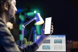Bearded man using cell phone with isolated white screen while using his laptop. Male freelancer holding smartphone with a blank chromakey template while working at his table. photo