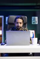 Young male freelancer sitting and working from home, utilizing his laptop for online research. Dedicated college student studying online, using modern technology to review information on internet. photo