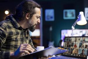 Bearded man with an annoyed expression, is holding a clipboard while having conference call with various people on his laptop. Male adult comparing documents with colleagues in a virtual meeting. photo