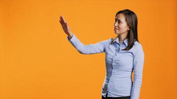 Asian woman examines holographic image reflection in studio, looking at modern artificial intelligence hologram over orange background. Confident person using metaverse projection. photo