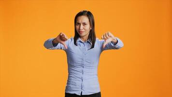 Asian model showing dislike symbol in studio, expressing disapproval and disagreement against orange background. Negative displeased girl giving thumbs down sign on camera. photo