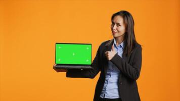 Business person holds laptop with greenscreen on camera, showing blank copyspace display on portable pc. Asian woman in formal suit presents computer with chromakey screen in studio. photo