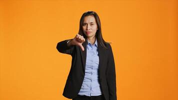 Asian female model giving dislike in front of camera, showing thumbs down with displeased facial expressions. Businesswoman does rejection and disapproval gesture over backdrop. photo