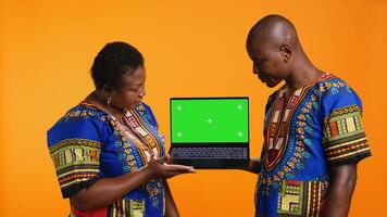 Ethnic couple showcasing laptop with greenscreen display, presenting isolated copyspace template and posing on camera. African american man and woman pointing at blank mockup layout. photo