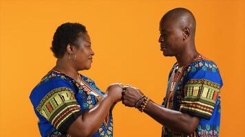 Playful ethnic couple doing fist bump gesture on camera, greeting each other with a cool modern sign in studio. Traditional african american people in colorful attire having fun over background. photo