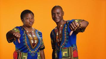 Ethnic man and woman showing thumbs down symbol over orange background, showcasing negative opinion with dislike sign. African american couple presenting disapproval and bad feedback. photo