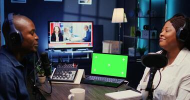 Green screen laptop next to vlogger show host and guest talking in neon lights ornate living room personal studio. Mockup notebook next to man and woman broadcasting discussion on online podcast photo