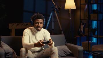 African american man in living room playing video games on gaming console, smashing controller buttons. Gamer wearing headphones, battling enemies in online multiplayer shooter from cozy apartment photo