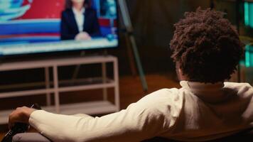 Man holding remote, watching news channel on television broadcast. Focus on person relaxing on couch in apartment by looking at widescreen smart TV set in blurry background photo