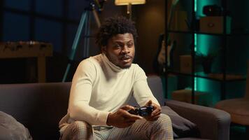 African american man sitting on couch at home, relaxing by playing videogames on gaming console. Gamer unwinding in apartment by competing in online multiplayer tournament photo