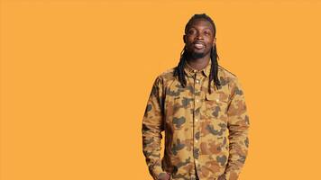 retrato de hombre con trenzas sonriente en frente de cámara, vistiendo frio camuflaje ropa y sensación contento en estudio. africano americano persona con de moda pelo posando terminado naranja fondo, relajado estilo. foto