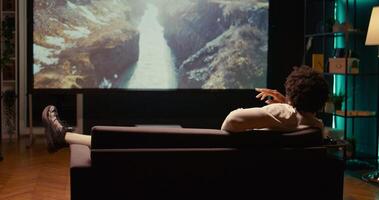 African american man watching nature documentary with beautiful cinematography on video projector. Person enjoying mountains and rivers scenery shots in home theatre, relaxing on couch photo