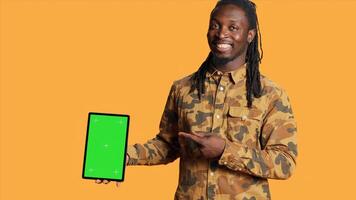 African american man presents greenscreen on tablet, showing isolated copyspace screen and smiling in front of camera. Young person holding mobile gadget with blank mockup display layout. photo